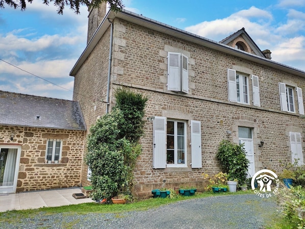 A stone building with white shutters, a small garden, and a gravel driveway.