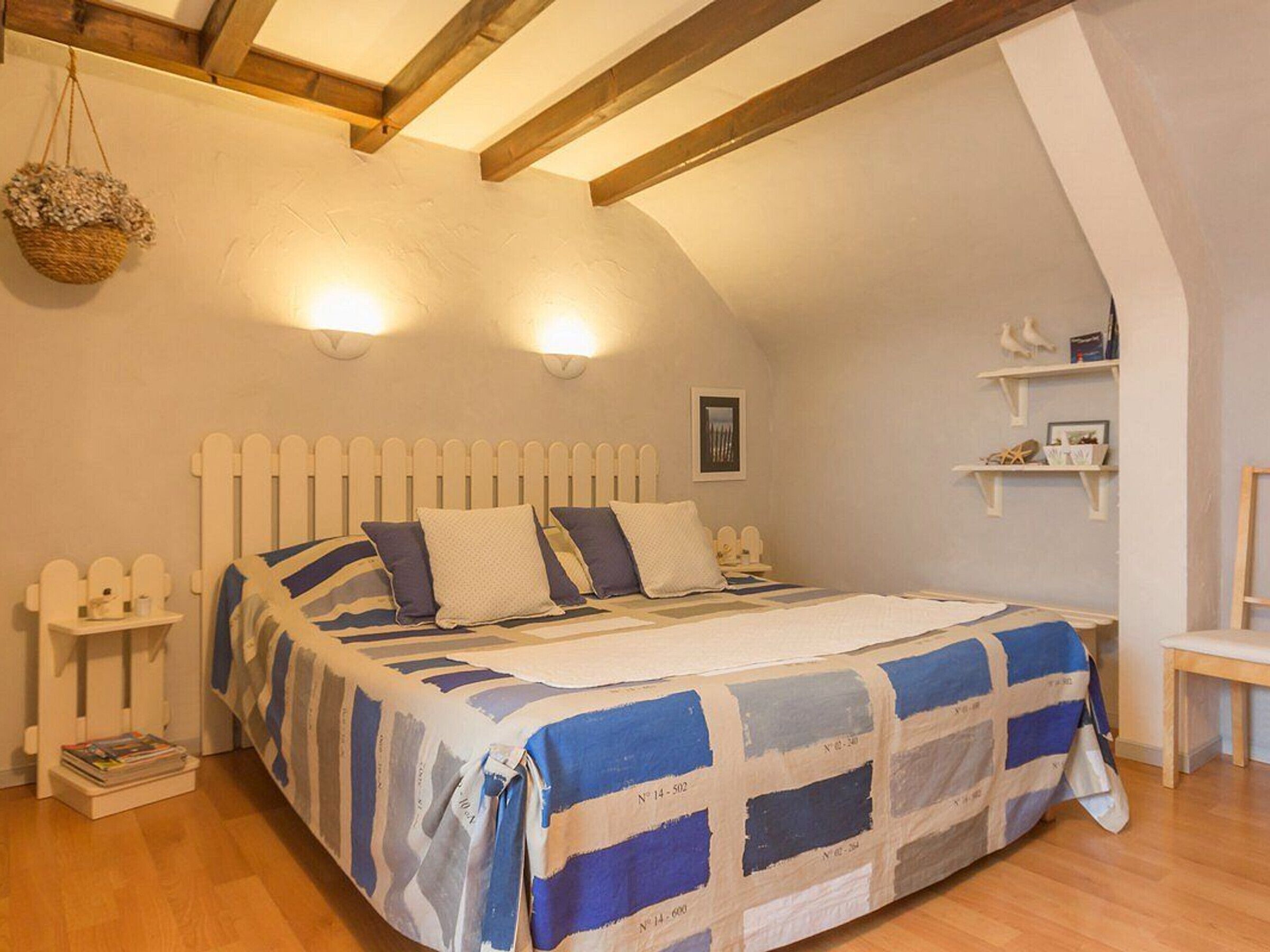 A bedroom with a wooden ceiling, a bed with a blue and white striped bedspread, a bedside table with a lamp, and a wall-mounted shelf with decorative items.