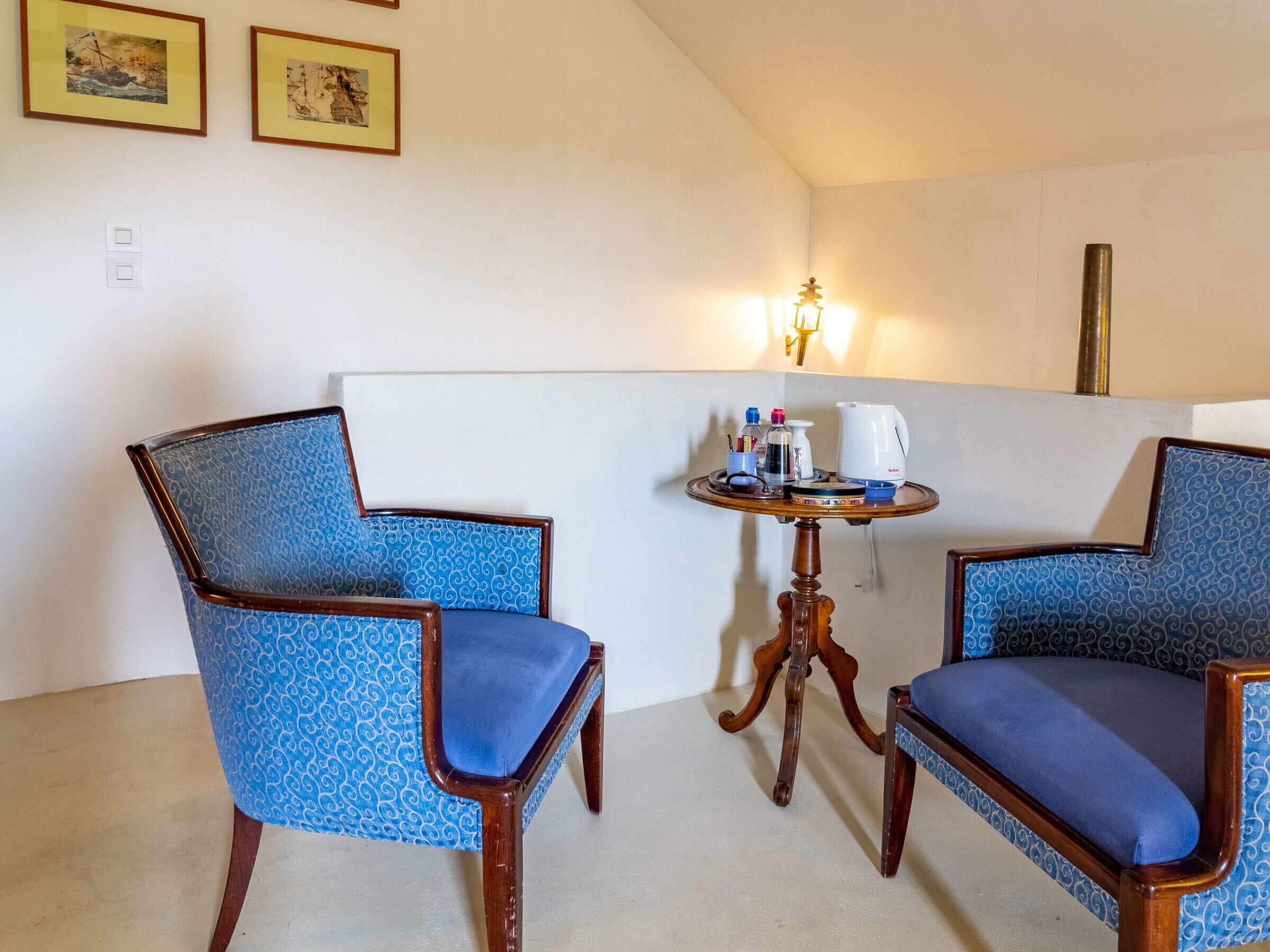 A room with two blue patterned chairs, a small round table with a coffee maker, and two framed pictures on the wall.