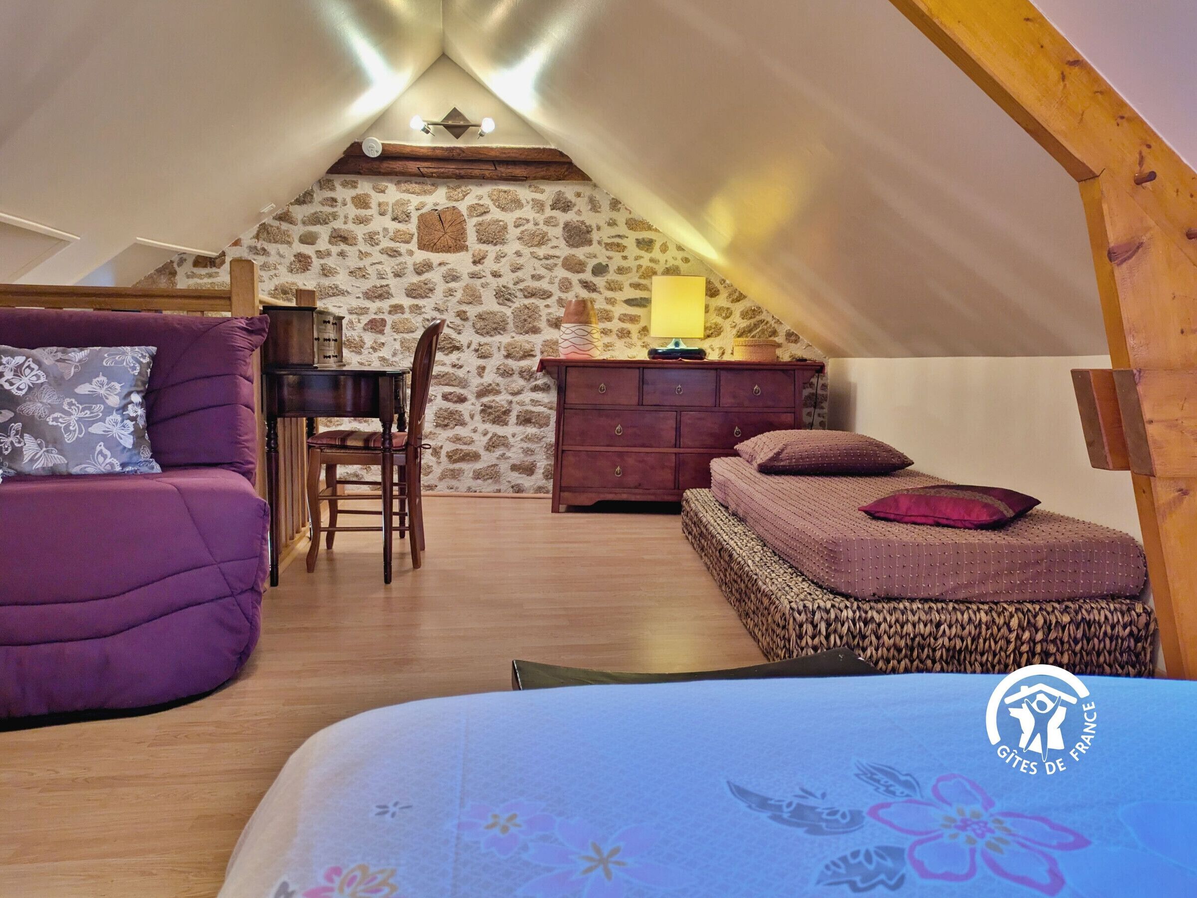 A cozy attic bedroom with a stone wall, a wooden desk, a purple sofa, and a bed with a patterned bedspread.
