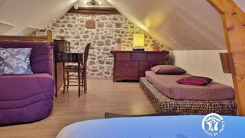 A cozy attic bedroom with a stone wall, a wooden desk, a purple sofa, and a bed with a patterned bedspread.