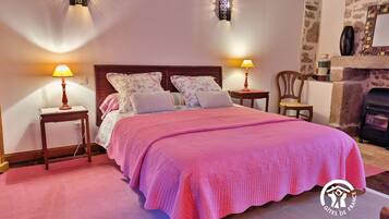 A bedroom with a large bed, two bedside lamps, a wooden headboard, a stone wall, and a fireplace.