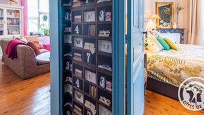 Una habitación con una amplia estantería llena de libros y secciones numeradas, un sofá acogedor, una cama con colcha estampada y una ventana con cortinas.