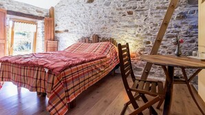 Un dormitorio con una cama de madera, una manta a cuadros, una silla de madera, una mesita pequeña y una pared de piedra.