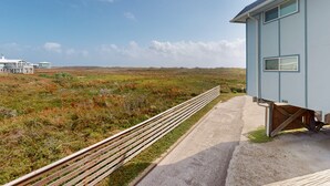 Une maison située sur la côte, avec un chemin en béton menant à une terrasse en bois qui offre une vue sur un paysage verdoyant.