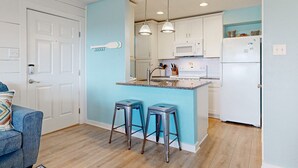 Une cuisine compacte dotée d’un îlot central, de meubles blancs et d’appareils électroménagers en acier inoxydable. L’îlot est agrémenté de deux tabourets, et un mur d’accent bleu apporte une touche de couleur.