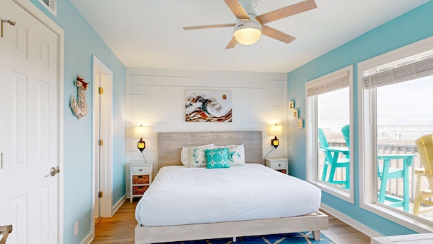 Une chambre à coucher avec un lit, un ventilateur de plafond, une grande fenêtre donnant sur un beau paysage et un mur bleu.