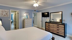 A bedroom with a bed, dresser, ceiling fan, TV, and artwork. - Villas on the Gulf #D3 | Quick Beach Access & Local Fun (Pensacola Beach)