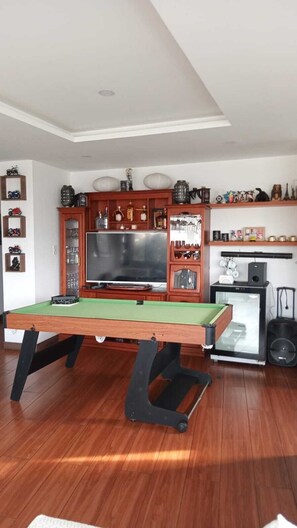 Una mesa de billar en una habitación con suelo de madera, un televisor y varios objetos decorativos en estantes.