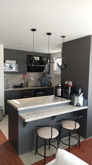 A modern kitchen with a central island, bar stools, hanging lights, and a window. - Departamento una Habitación con Jacuzzi (Quito)