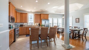 A spacious kitchen with wooden cabinets, a central island, and a dining area with wicker chairs. - The Beachcomber - Nags Head MP 12, 125 Yds to Beach, Ocean View, Elevator, Pool, Hot Tub (Nags Head)