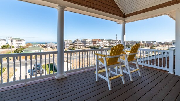 A wooden deck with white railings, two yellow Adirondack chairs, and a view of a beach and houses in the background. - The Beachcomber - Nags Head MP 12, 125 Yds to Beach, Ocean View, Elevator, Pool, Hot Tub (Nags Head)