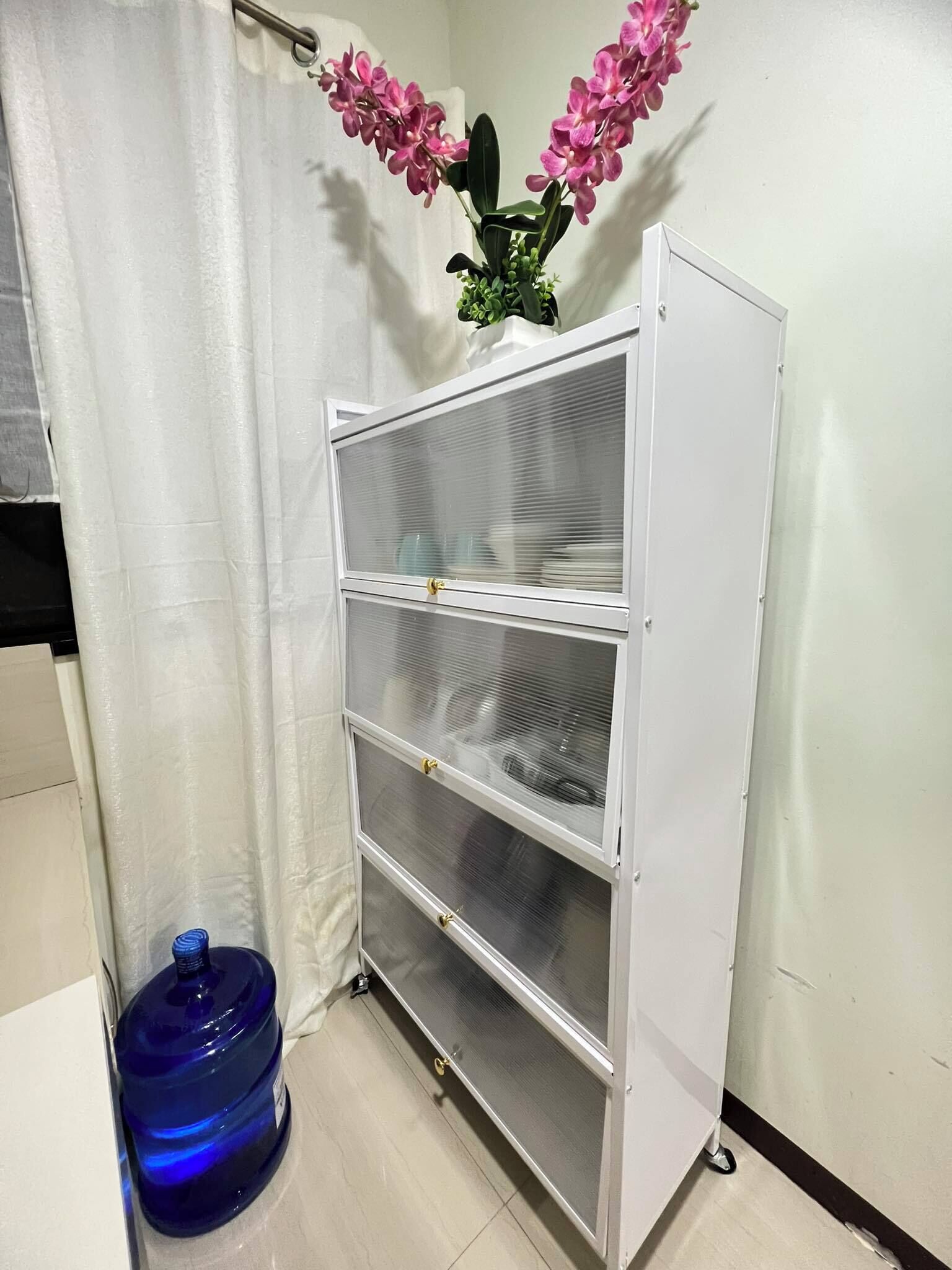 A white metal shelving unit with three glass doors, a blue water dispenser, and pink flowers on top.
