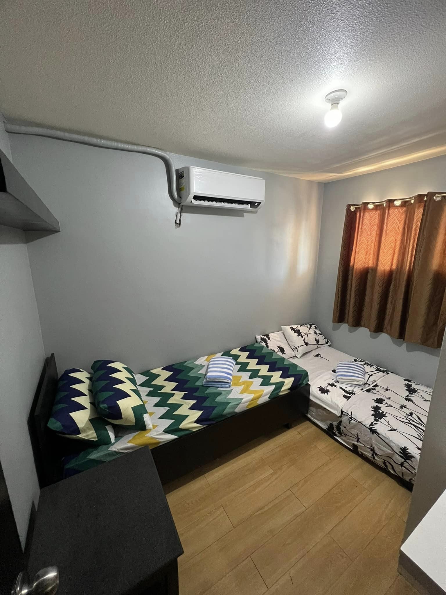 A compact room with a bed, a small desk, and an air conditioning unit mounted on the wall.
