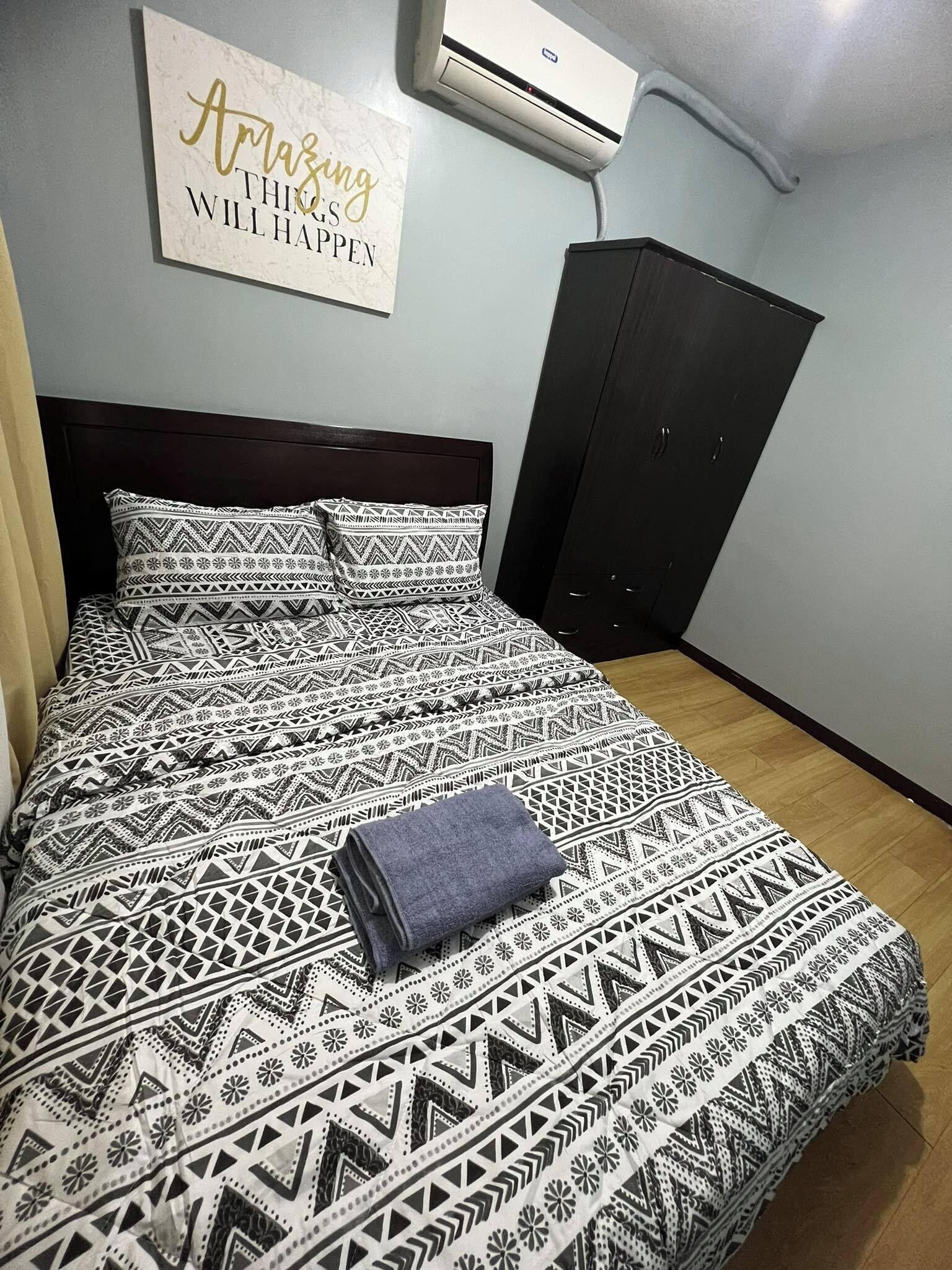 A neatly made bed with patterned bedding, a wall-mounted air conditioner, and a motivational quote.