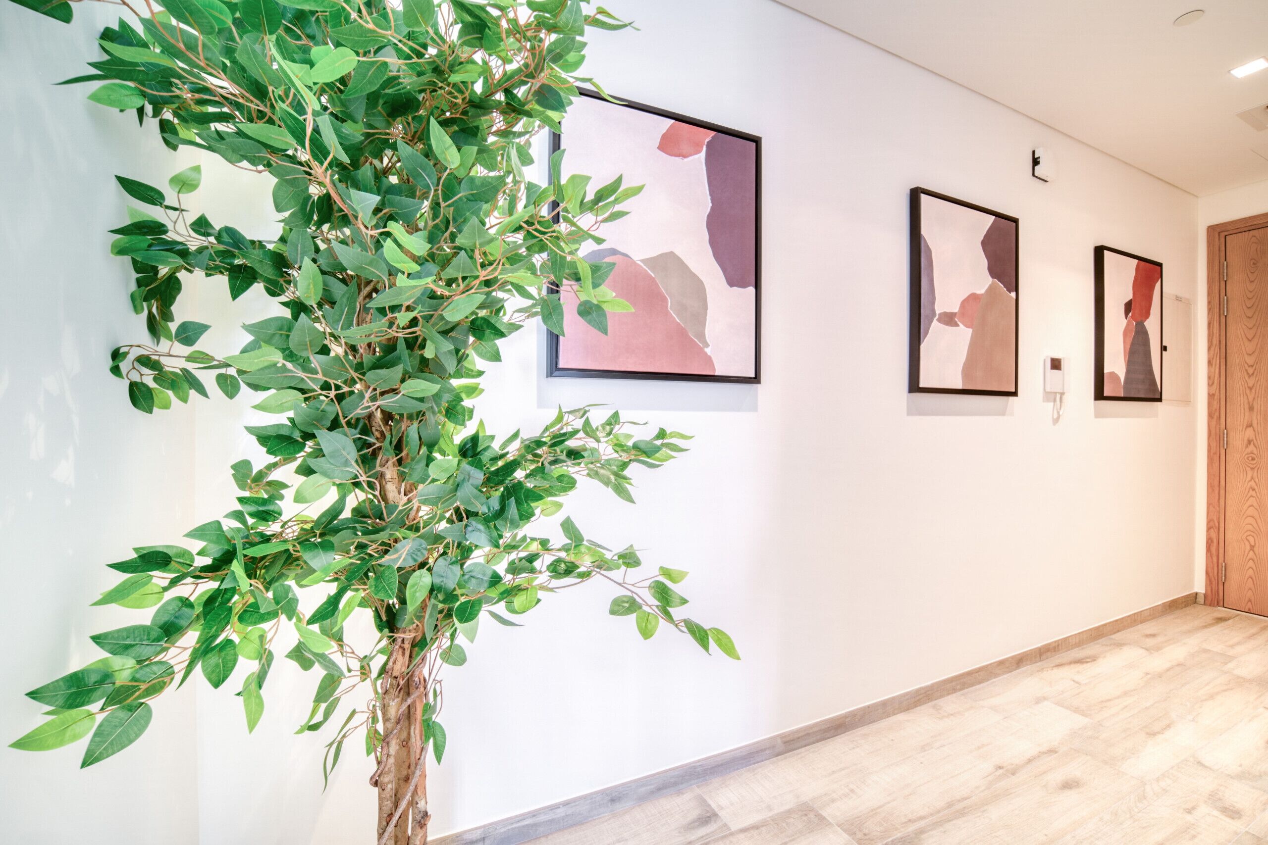 A hallway with a large artificial tree, framed artwork on the walls, and a wooden door at the end.