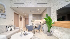 A modern living room with a sofa, coffee table, and a dining area with chairs. There is a kitchen area with a sink and a large potted plant. - Serene 1BR Apartment in Azizi Riviera 27, Al Merkadh by Deluxe Holiday Homes (Dubai)