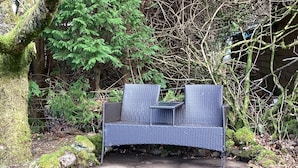 A wicker bench with a backrest and armrests, positioned on a stone pathway surrounded by greenery and trees. - Taigh Sealladh Fèidh B&B (Invergarry)