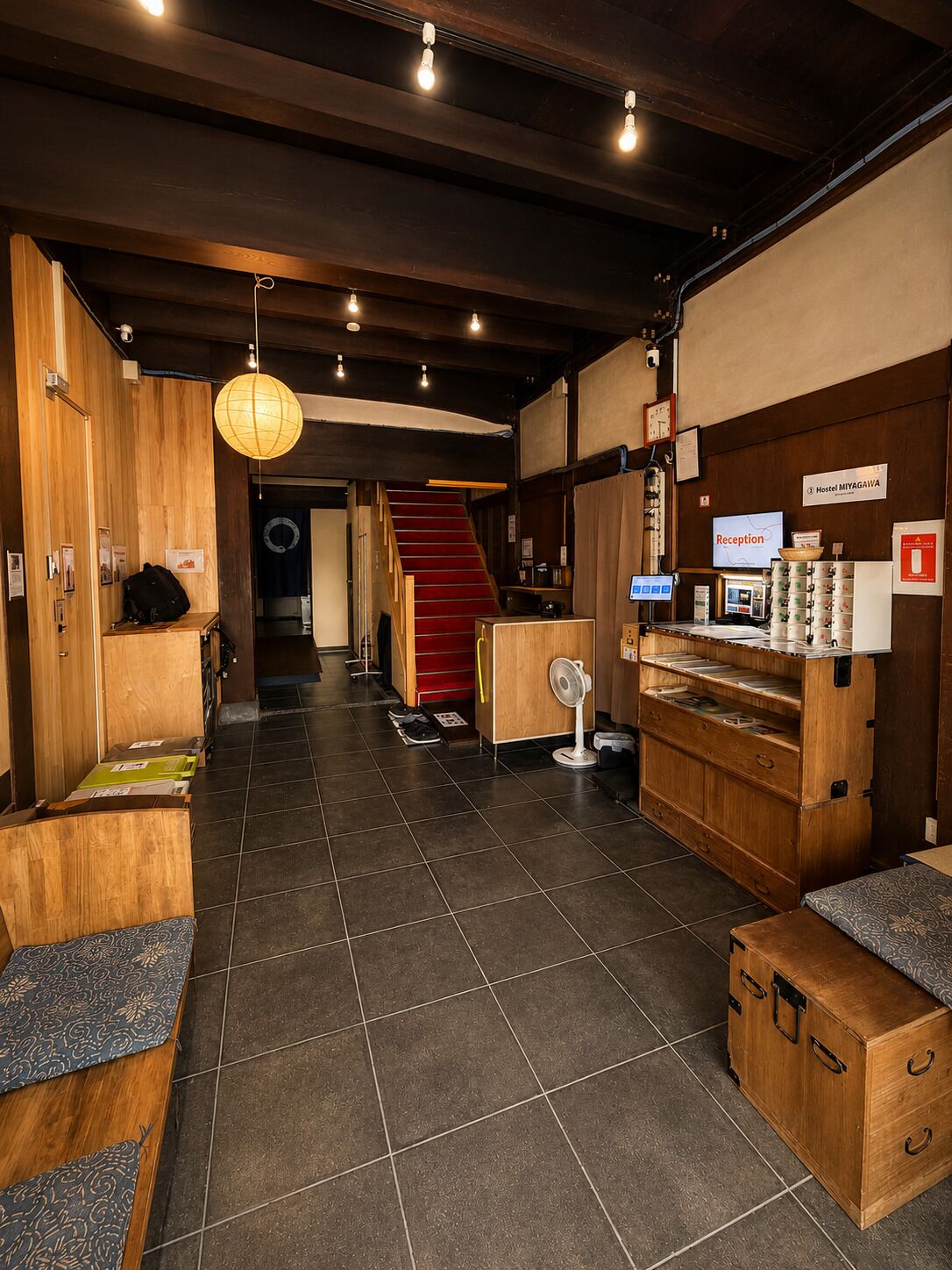 A reception area with a reception desk, a clock, a sign for 'Hostel MIYAGAWA,' and a staircase leading to another floor.