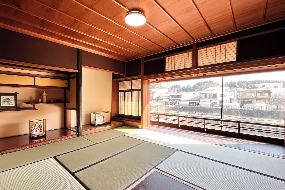 Ett traditionellt japanskt rum med tatamimattor på golvet, ett lågt trätak och utsikt över en stad genom stora fönster.