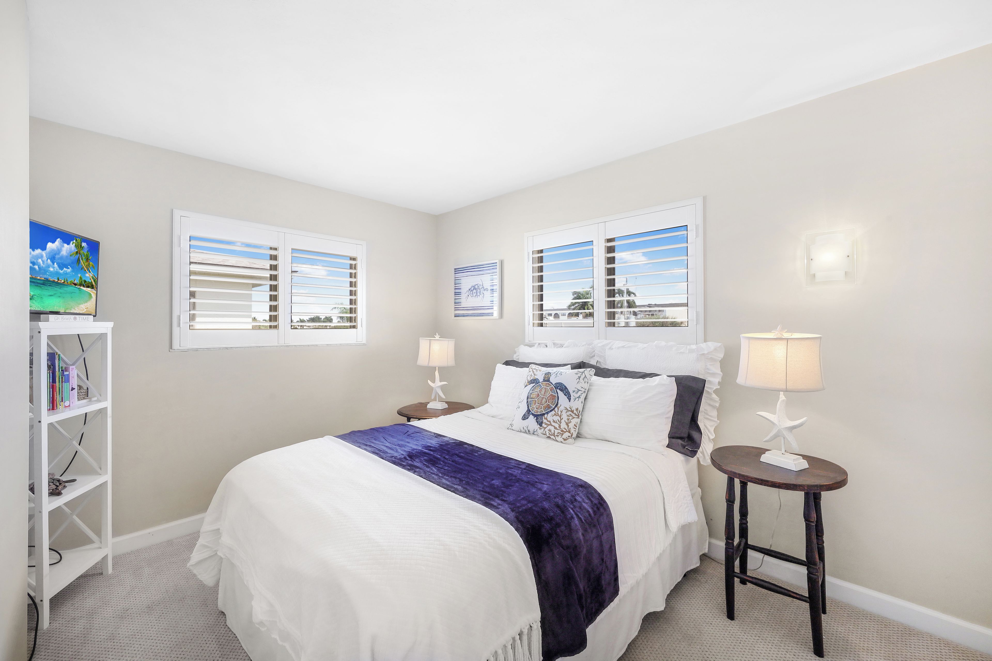 A bedroom with a bed, two bedside tables, a bookshelf, and two windows with blinds.