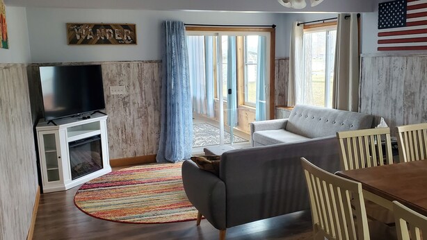 A living room with a TV, sofa, dining table, and chairs. There is a large window with curtains, a wooden cabinet, and a flag on the wall. - Hide-a-way at Swan Lake: fishing, relaxing, and dog friendly. (Allegan)