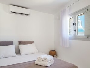 A hotel room with a bed, pillows, a rolled towel, an air conditioning unit, a window with curtains, and a view of a building outside. - MyMykonos Guest House  (greece)