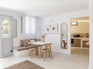 Uma sala de estar moderna com um sofá, uma mesa de jantar redonda e cadeiras. Há uma janela com cortinas leves, um espelho fixado na parede e uma pequena área de cozinha visível por uma porta aberta.