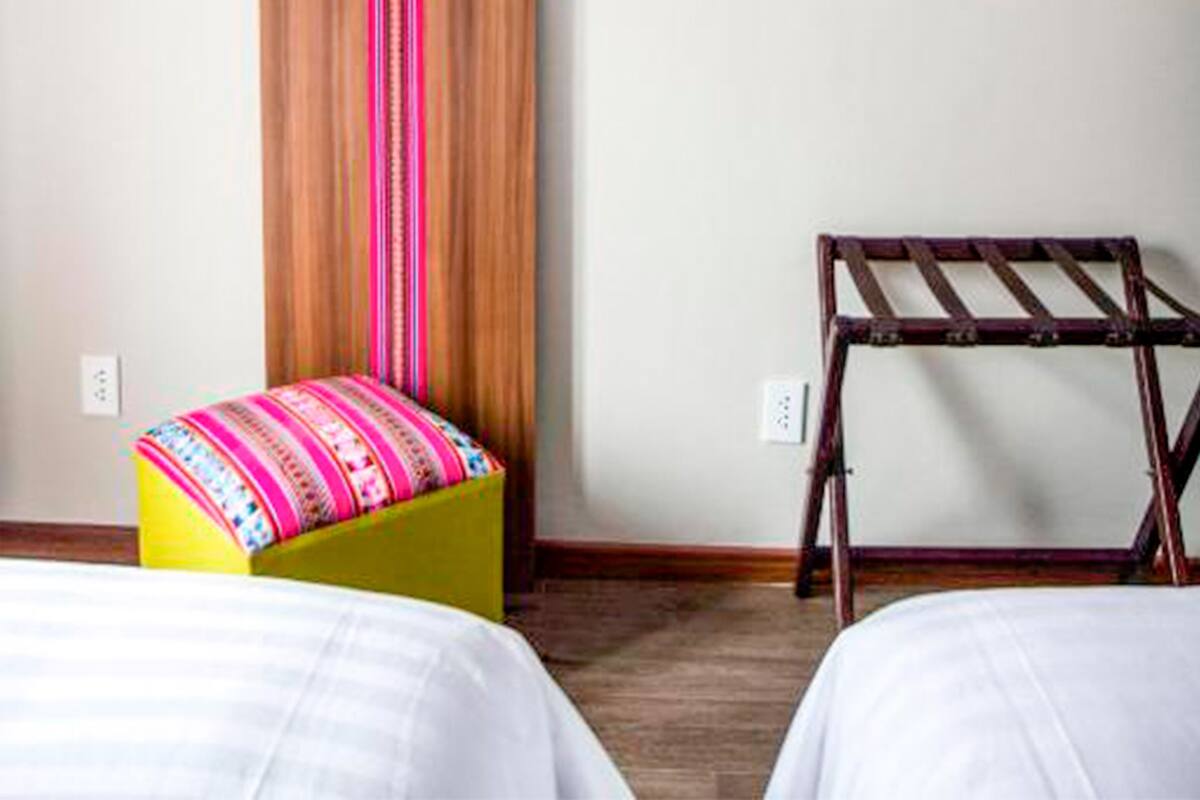 Een hotelkamer met twee bedden, een flatscreen televisie aan de muur bevestigd, een houten paneel met een verticaal gestreept patroon, een kleurrijk poufje en een houten stoel.