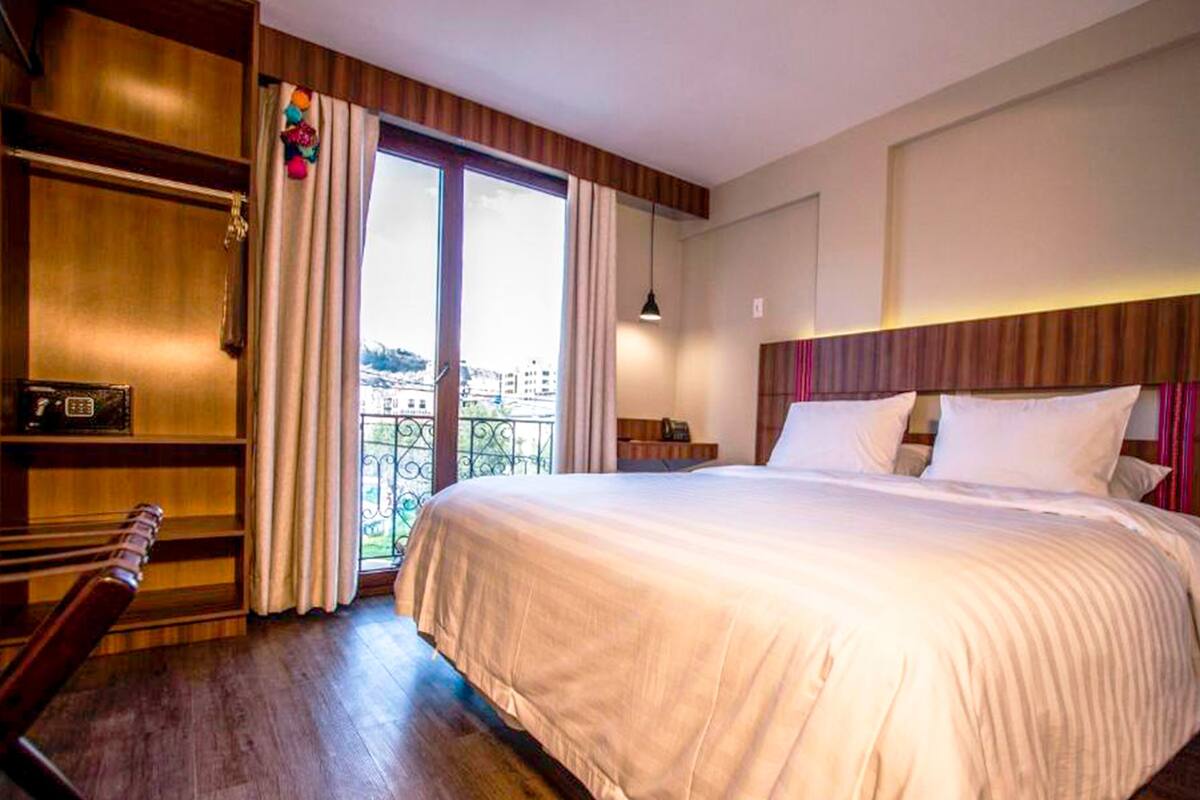 Een hotelkamer met een groot bed, houten vloer, een bureau met stoel, een nachtkastje en uitzicht op de stad door het raam.