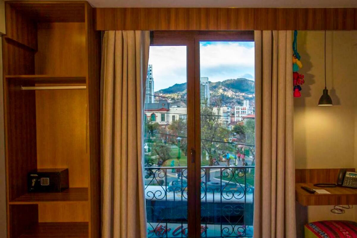 Een hotelkamer met een houten kast, een bed, een bureau met telefoon en uitzicht op een stadsbeeld door een groot raam met gordijnen.