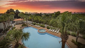 Une piscine entourée de bâtiments, avec des chaises longues et des palmiers, le tout baigné par la lumière d’un coucher de soleil.