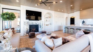 A modern living room with a fireplace, a large TV, and a kitchen area with bar stools. - Desert Sapphire at Desert Color (by Ember Stays) (St. George)
