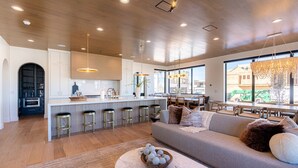 A modern kitchen with a central island, bar stools, and a dining area with a large window. - Desert Sapphire at Desert Color (by Ember Stays) (St. George)