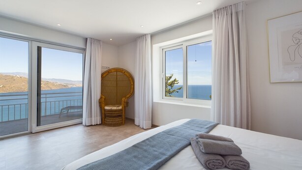 A bedroom with a large bed, a view of the sea, a wicker chair, and a window with curtains. - Bienvenido in der Villa Los Pinos (Velilla-Taramay)