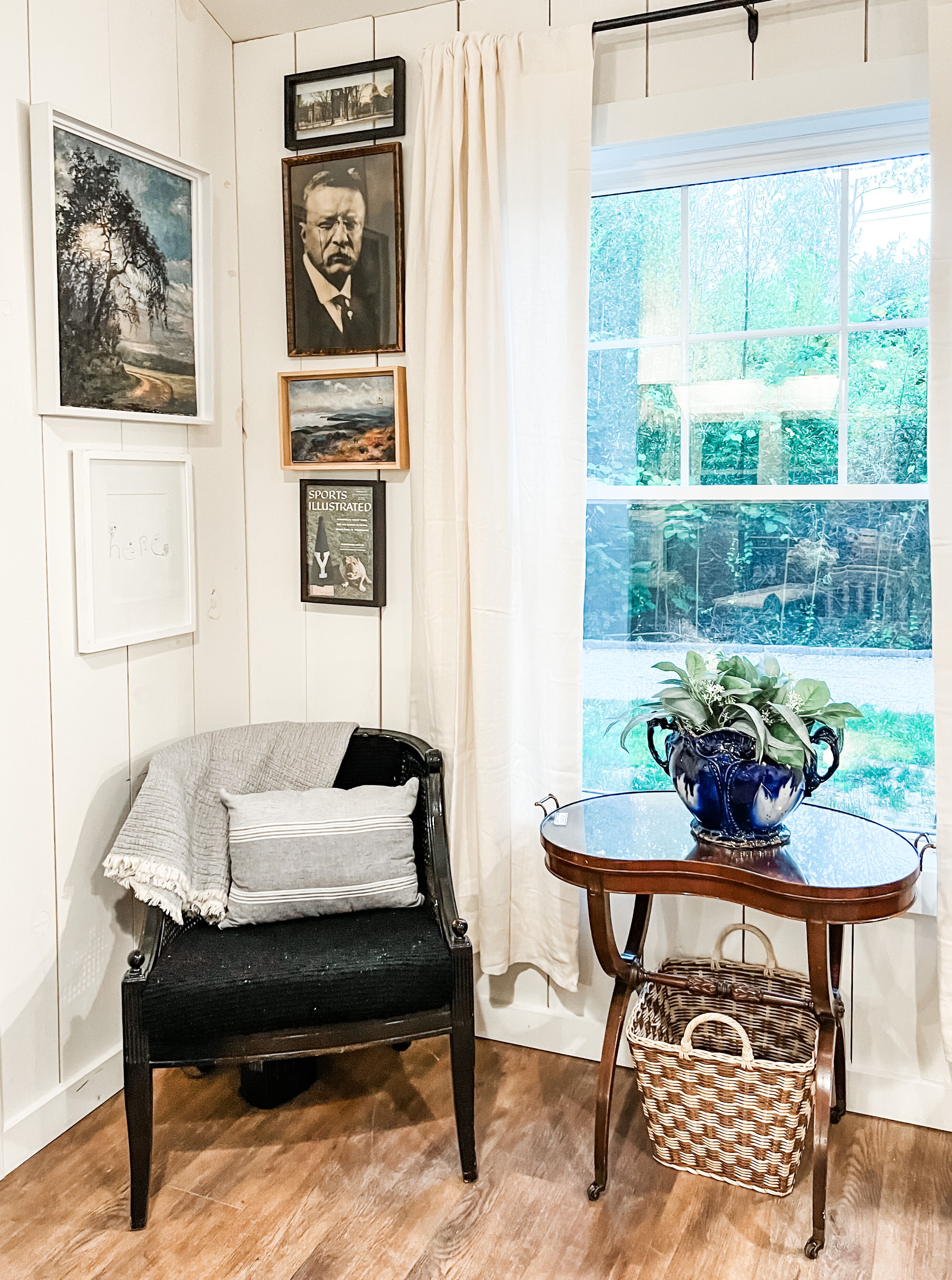 A room with a wooden floor, a black chair with a grey throw, a small table with a blue vase, and a window with a view of greenery.