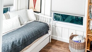 Ein Schlafzimmer mit einem Bett, einem gerahmten Bild an der Wand, einem Fenster mit Jalousien und einem Korbwäscher.