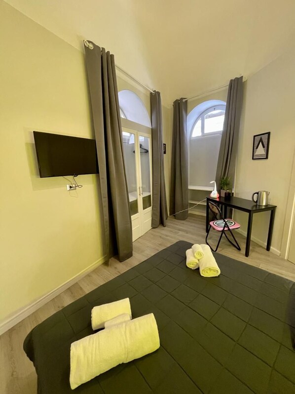 A room with a bed, a TV mounted on the wall, a small table with a kettle, and a chair.