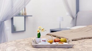 En pænt anrettet bakke med morgenmad på sengen, med et blondehæftet sengetæppe, en lille urtepotte med en orchidé, et glas appelsinjuice, en kop med et logo og en tallerken med frugt og et wienerbrød.
