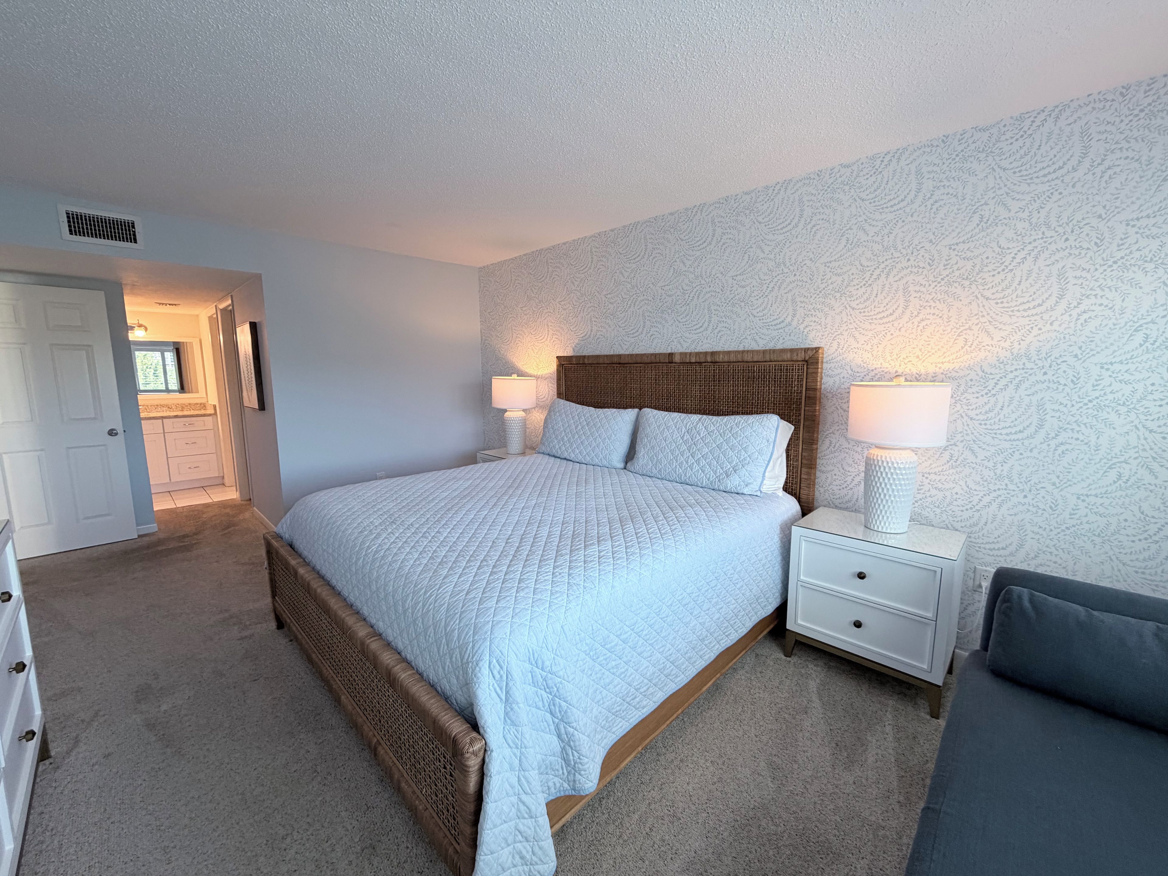 A bedroom with a bed, bedside tables, a nightstand, a chair, and a bathroom visible through an open door.