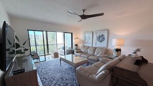 Un salon comprenant un canapé, une table basse et un téléviseur. On y trouve également un ventilateur de plafond et une grande fenêtre donnant sur un paysage verdoyant.