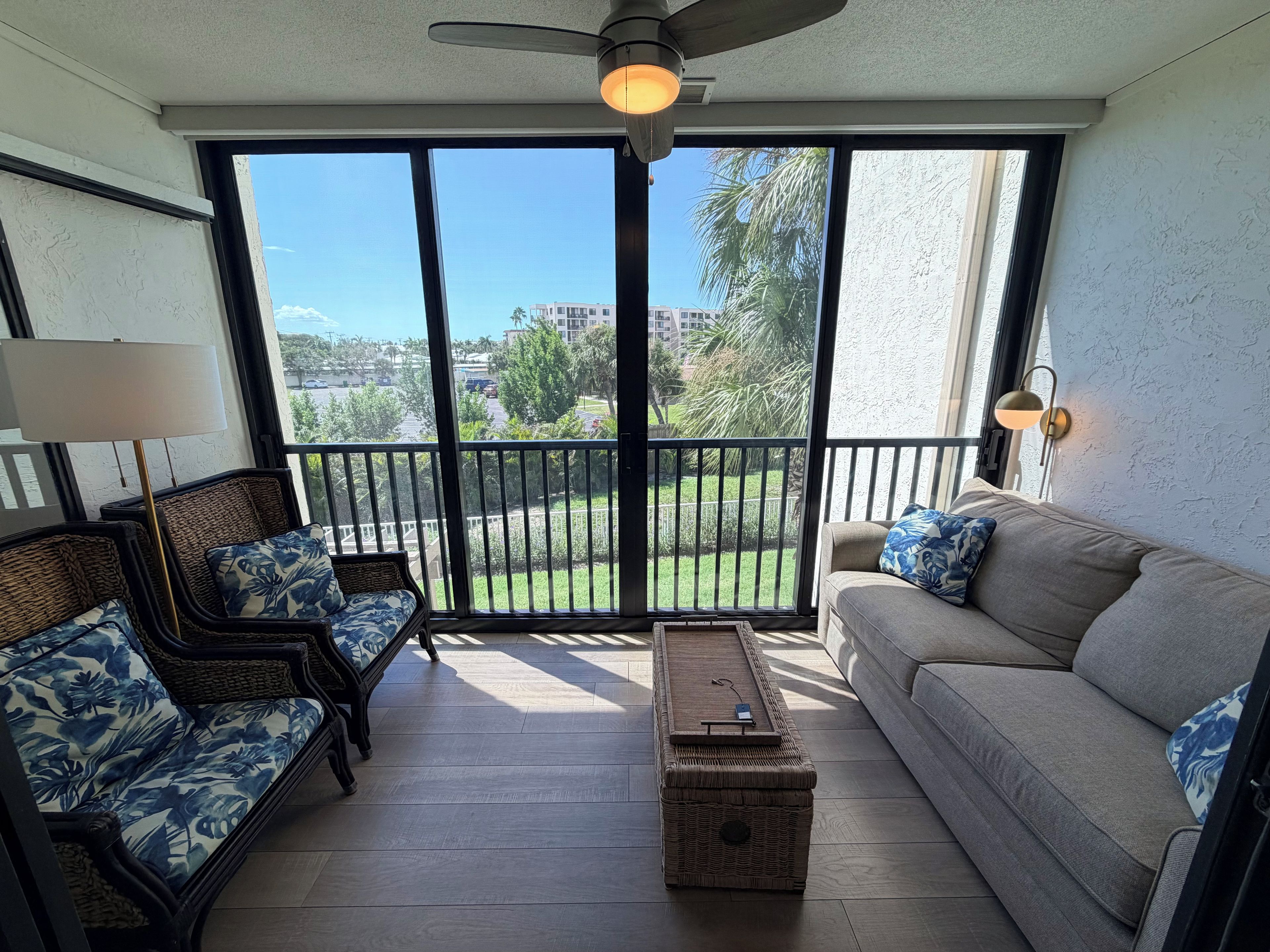 A balcony with a ceiling fan, a sofa, two wicker chairs, a coffee table, and a view of a green landscape.