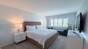 Une chambre à coucher comprenant un lit, des tables de chevet, une commode, une fenêtre avec des stores et un téléviseur.