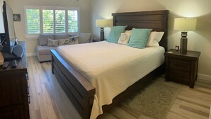 A bedroom with a large bed, two bedside tables with lamps, a wooden headboard, a window with blinds, a sofa with cushions, and a television on a stand. - Recently renovated third floor (Sarasota)