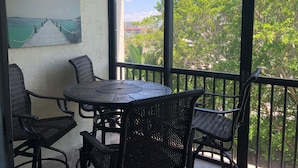 A balcony with a round table and four wicker chairs, a painting of a pier, and a view of trees and buildings. - Recently renovated third floor (Sarasota)