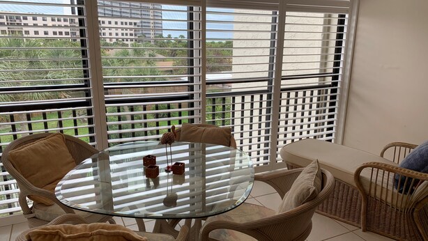 Uma varanda com móveis de vime, uma mesa de vidro e vista para edifícios e áreas verdes.