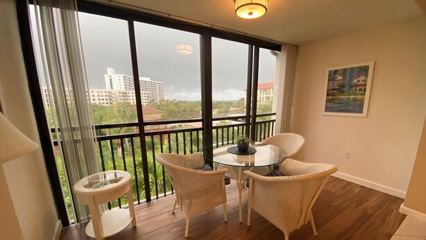 Een balkon met rieten meubels, een glazen tafel en uitzicht op gebouwen en groen.