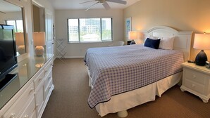 Een slaapkamer met een bed, een televisie, een plafondventilator, een raam met jaloezieën en een lamp op het nachtkastje.