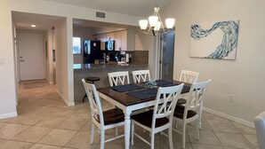 A dining area with a dark wooden table and white chairs, a chandelier, and a kitchen in the background. - Bright and spacious 3 bedroom, 4th Floor unit (Sarasota)