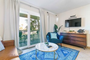 A living room with a sofa, a coffee table, a blue armchair, a TV mounted on the wall, and a sliding glass door leading to an outdoor area with chairs.
